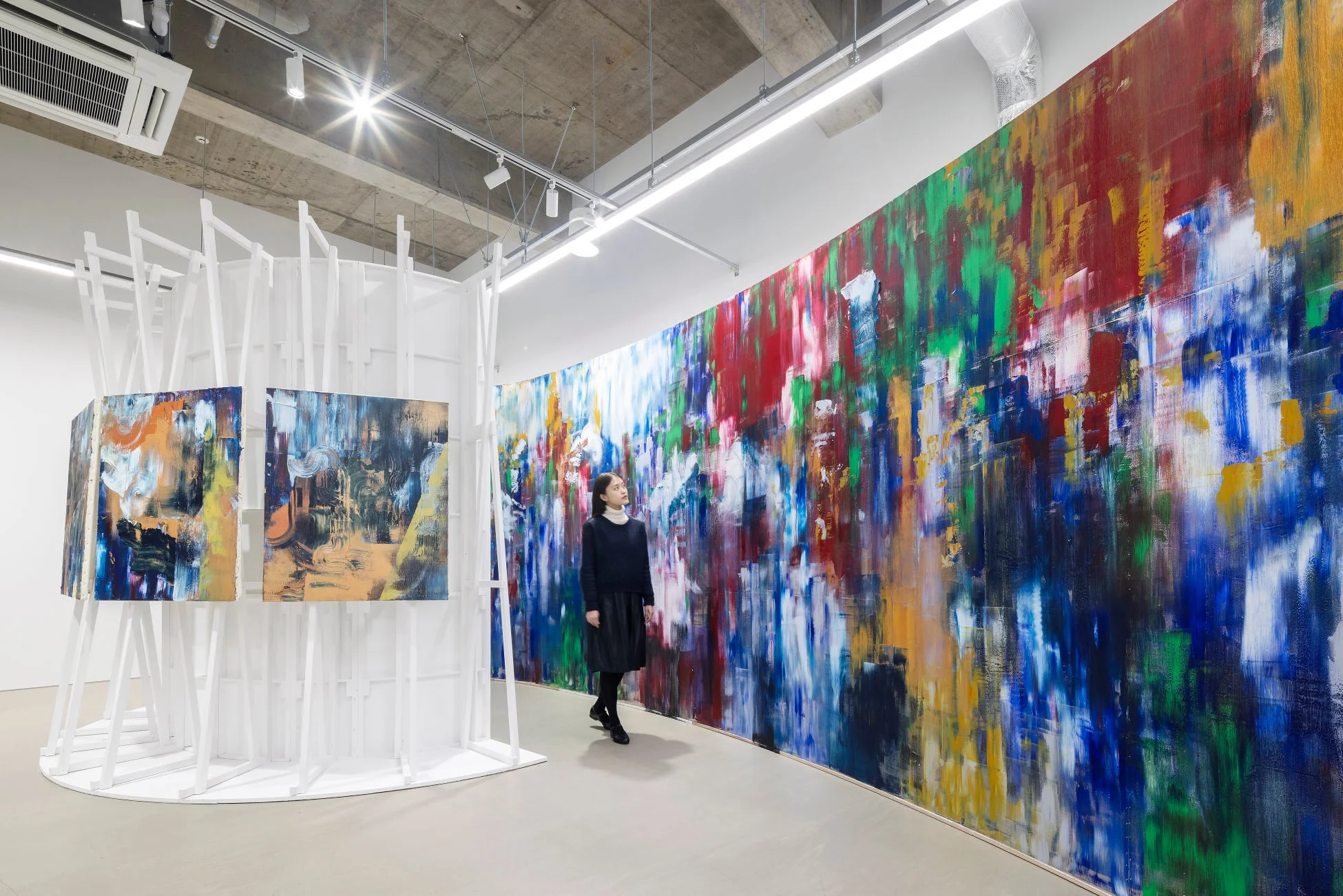 現代アートの展示空間で、一人の女性が鮮やかな色彩の抽象画が描かれた大きな壁画を鑑賞している