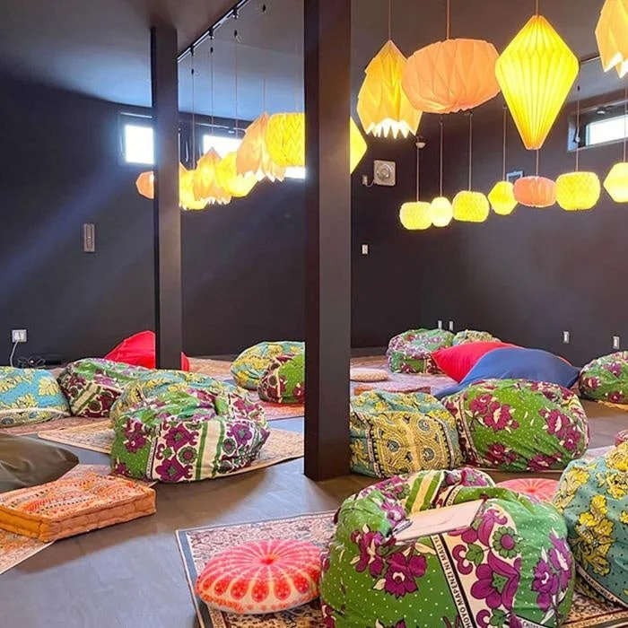 暗い壁の部屋に、様々な形をした黄色やオレンジ色の提灯が吊るされ、温かい光を放っています。床には柄物のラグが敷かれ、カラフルなビーズクッションやフロアクッションが多数配置されており、リラックスできる空間を演出しています。