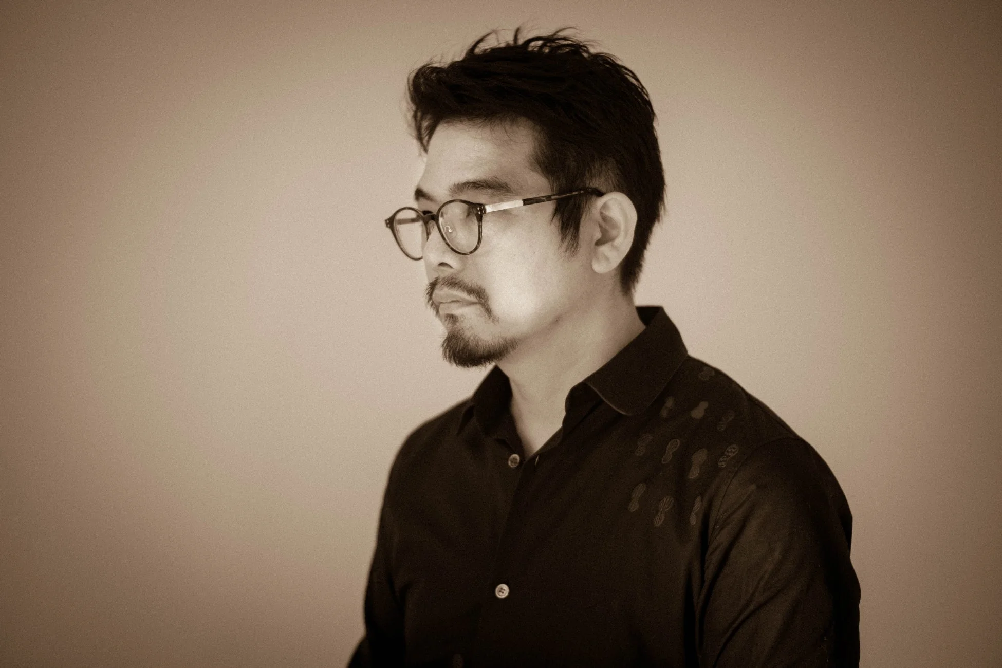 眼鏡とヒゲを蓄えた男性がセピア調の背景を背に左を向いているポートレート。黒いシャツを着用し、落ち着いた雰囲気で思索にふけっているような表情です。