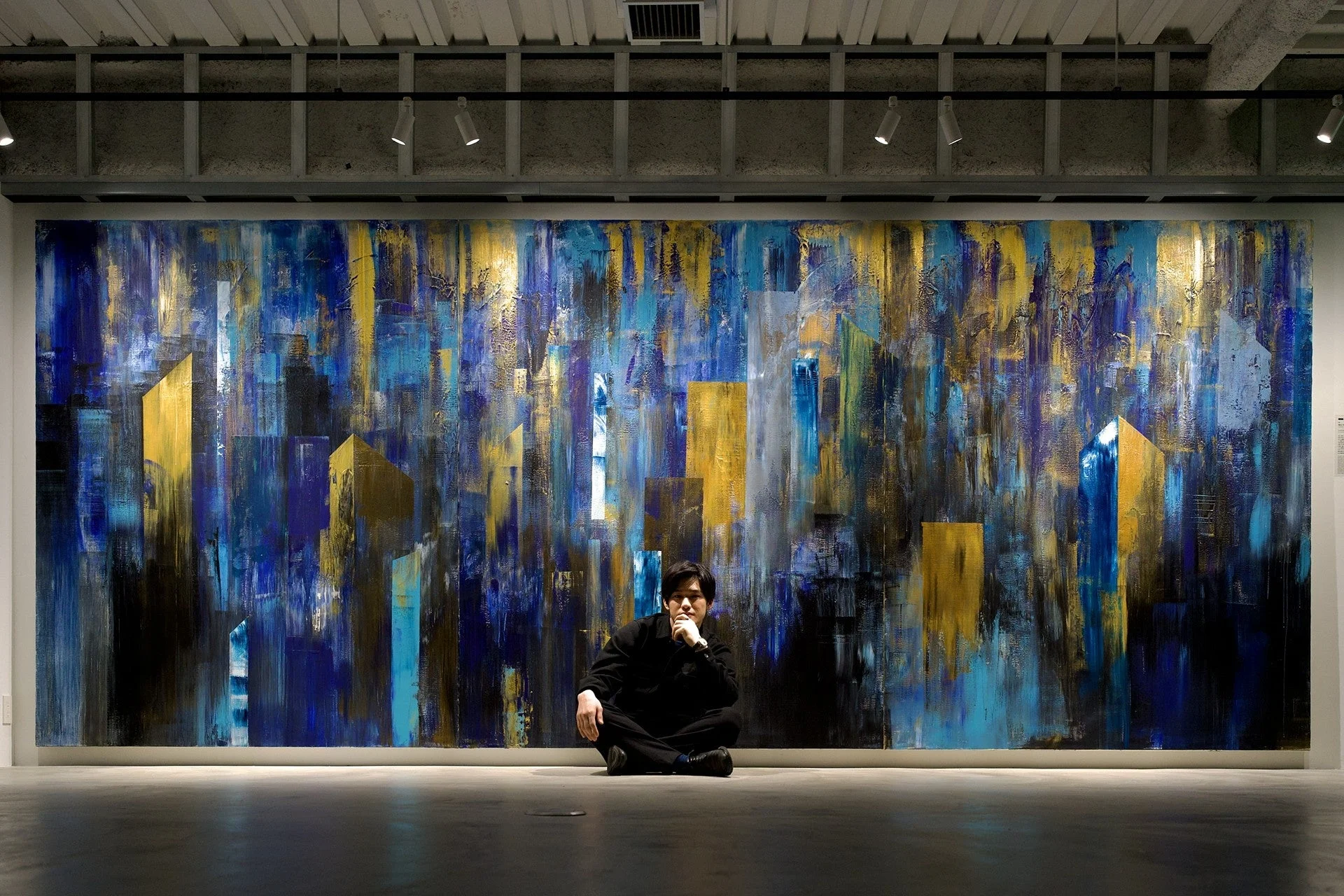 広々としたギャラリー空間で、青と金色を基調とした巨大な抽象画の前に一人の男性が座っている