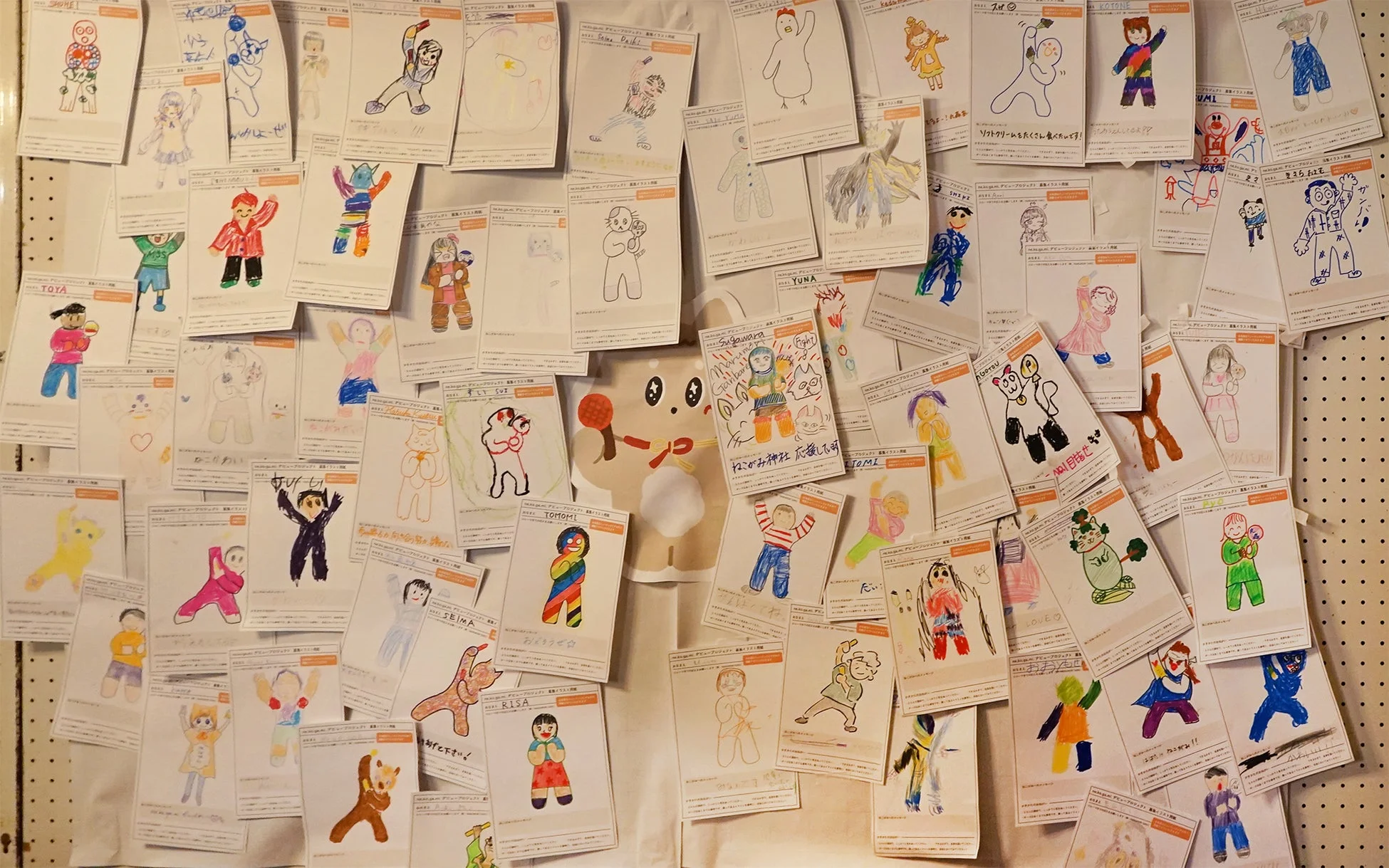壁一面に、様々な色と形で描かれた無数の子供たちのイラストが貼られています。多くは人や動物のキャラクターで、名前や応援メッセージなどの日本語の文字も見られます。