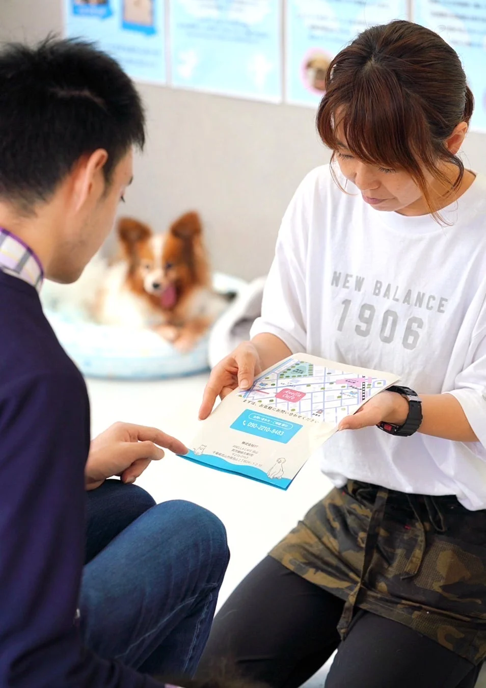 男性と女性がパンフレットを見ながら話し合っており、背景には犬が写っています。ペット関連のサービスについて相談している様子です。