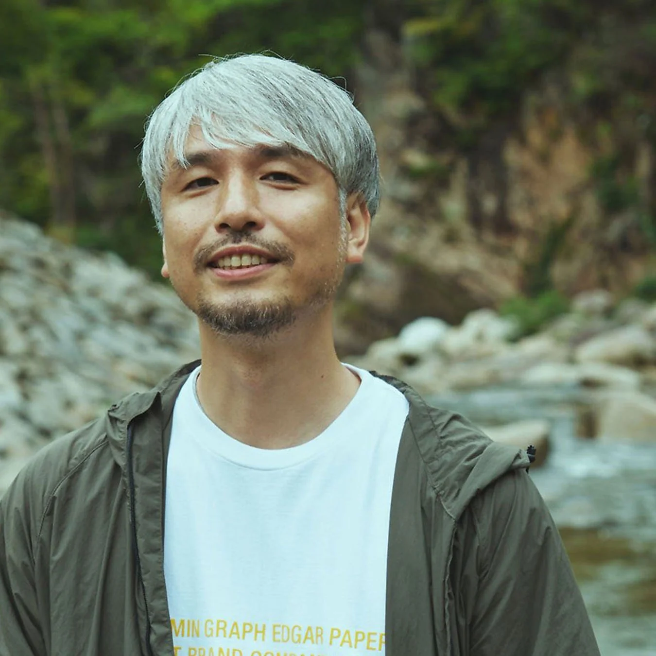 白髪と髭が特徴の男性が、自然豊かな川辺で笑顔を見せています。