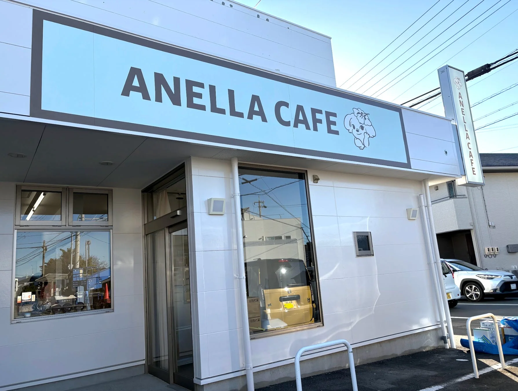 白いモダンな建物の「ANELLA CAFE」の外観です。大きな看板には店名と可愛らしい犬のロゴが描かれており、青空の下、明るい雰囲気のカフェであることが分かります。