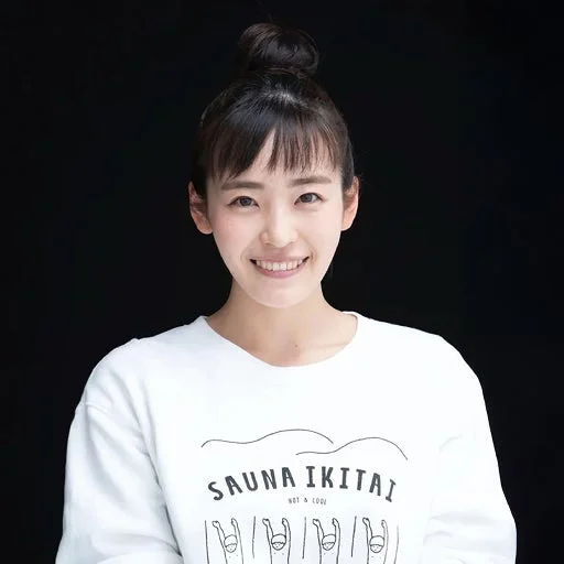 お団子ヘアの若い日本人女性がカメラに向かって笑顔を見せています。白いスウェットシャツには「SAUNA IKITAI」という文字とシンプルなイラストが描かれています。