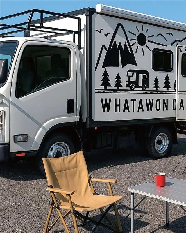 キャンピングカーの横にキャンプ用の椅子とテーブルが置かれ、リラックスしたアウトドアの雰囲気を醸し出しています。車両には山や木、太陽のデザインが施されています。
