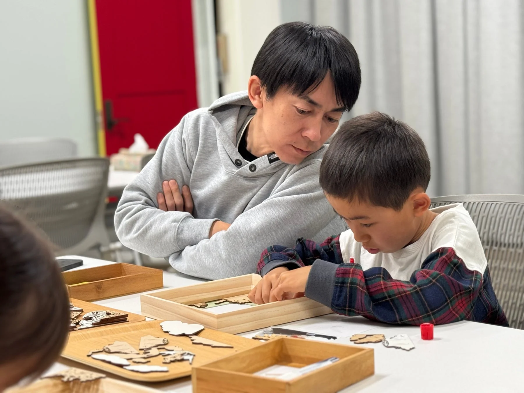 テーブルで木製のパズルか工作に集中する男の子と、それを見守る男性