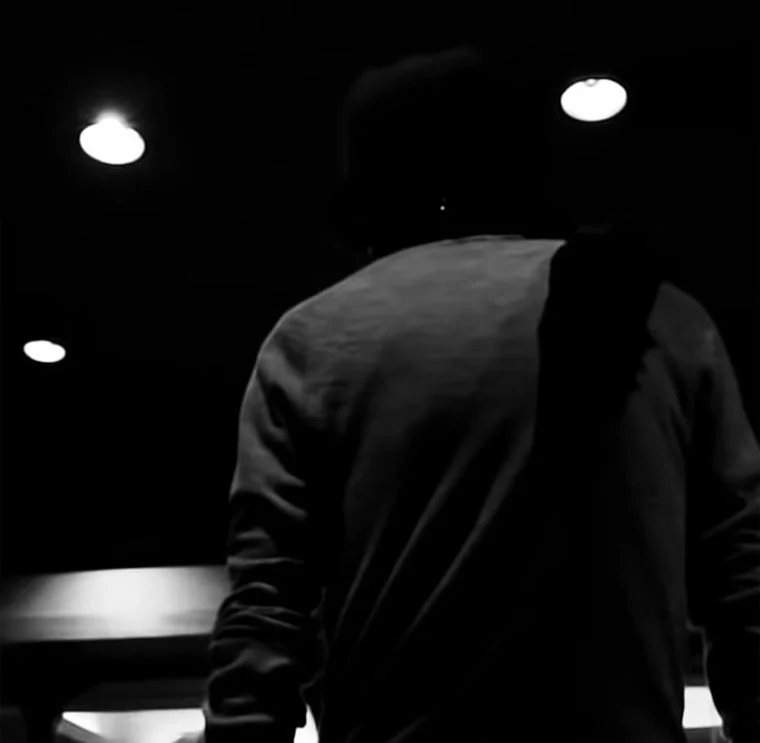 暗い空間で、上部に複数の光源がある中、一人の人物が後ろ姿で立っているモノクロームの写真。