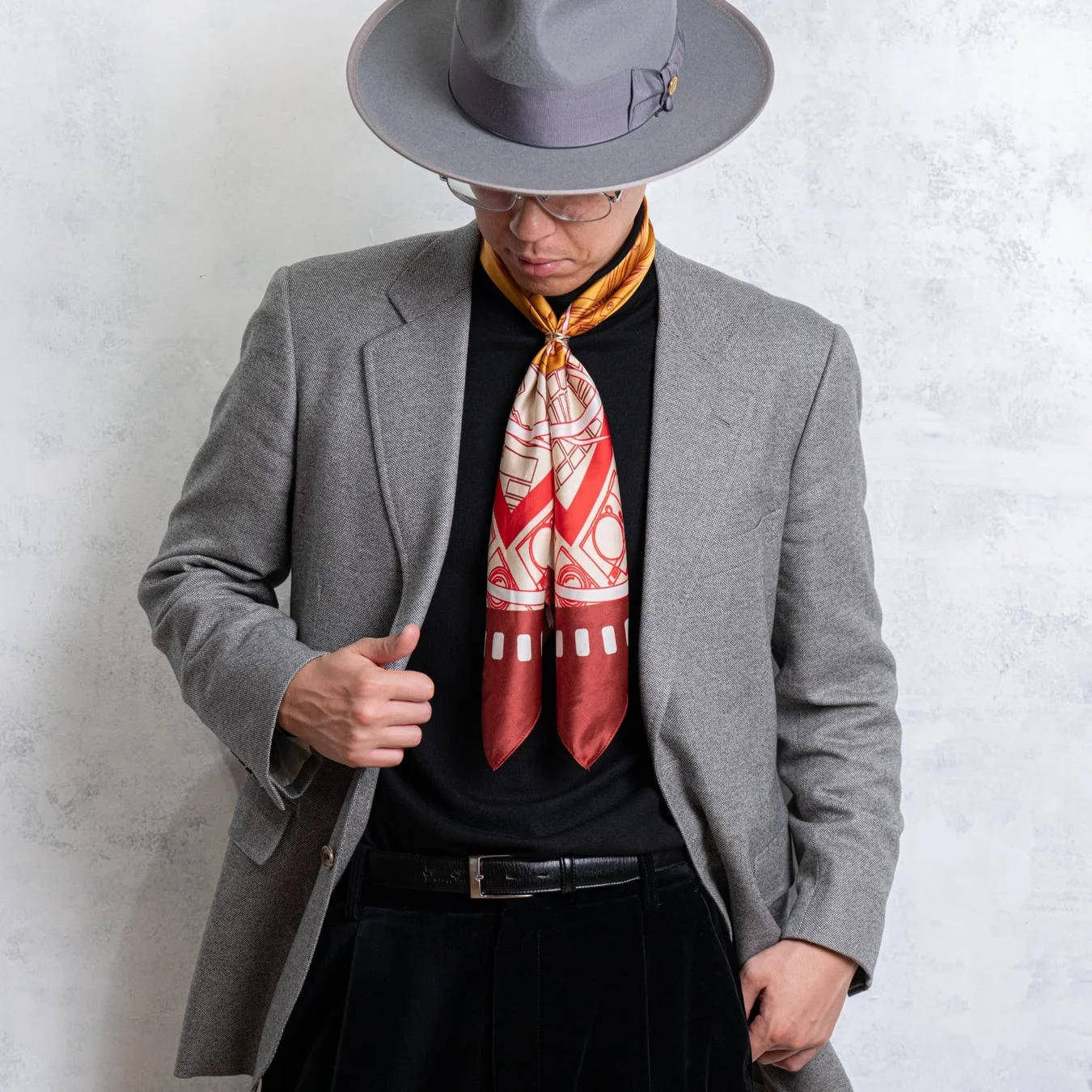 グレーのフェドラハットとジャケットを着用し、黒のタートルネックに赤い柄のスカーフを合わせた男性のスタイリッシュな上半身のポートレート。落ち着いた色合いの中にスカーフがアクセントになっています。