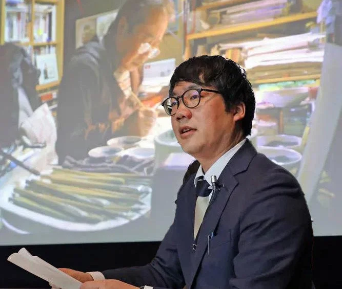 眼鏡をかけたスーツ姿の男性が、資料を手に講演またはプレゼンテーションを行っています。背景のスクリーンには、職人が繊細な作業をしている映像が映し出されており、何らかの専門的な内容について話していることが伺えます。