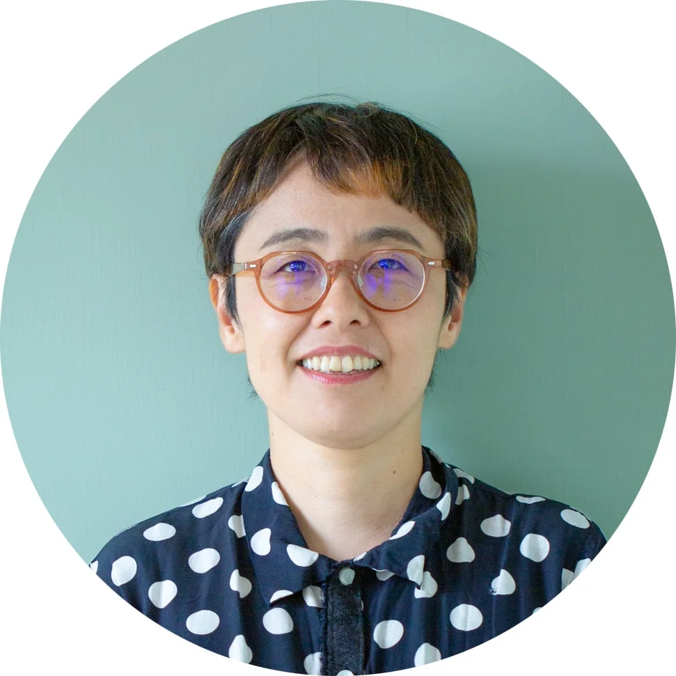 水色の背景の前で、眼鏡をかけ、ショートヘアの女性が笑顔でカメラを見つめているポートレートです。水玉模様のシャツを着用しています。