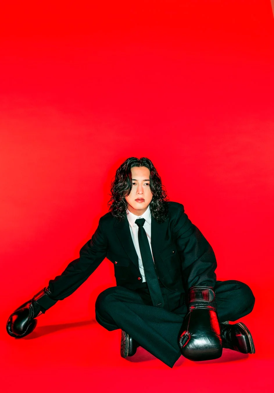 鮮やかな赤い背景の前で、黒いスーツとボクシンググローブを着用し、長いカールヘアの男性が座っているポートレート写真
