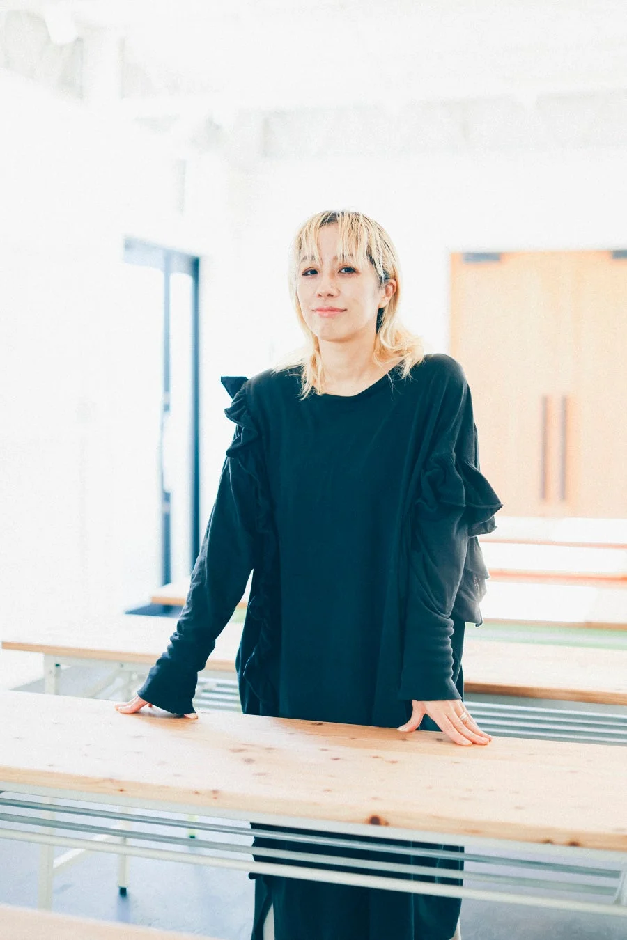 金髪の若い女性が、黒いフリルのトップスを着て、明るい屋内の空間で木製のテーブルに寄りかかっています。