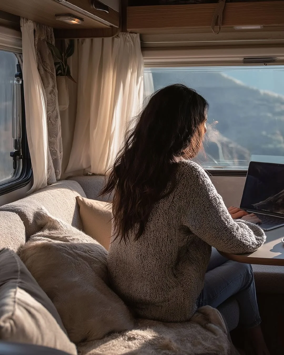 キャンピングカーの窓辺で、女性がノートパソコンを使って作業しています。窓の外には美しい山の景色が広がり、くつろいだ雰囲気の中でリモートワークをしている様子が伺えます。