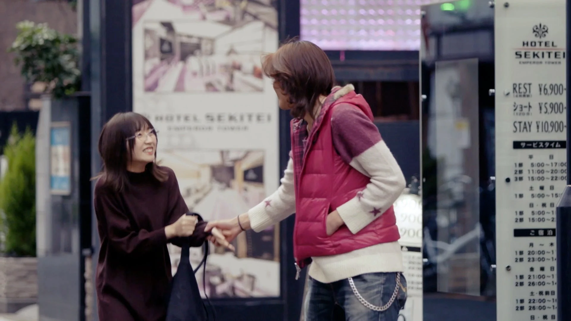 街中で二人の人物が交流している。女性は笑顔で、男性から何かを受け取っているようだ。背景には「HOTEL SEKITEI」の料金表が見え、都会の日常の一コマを捉えている。