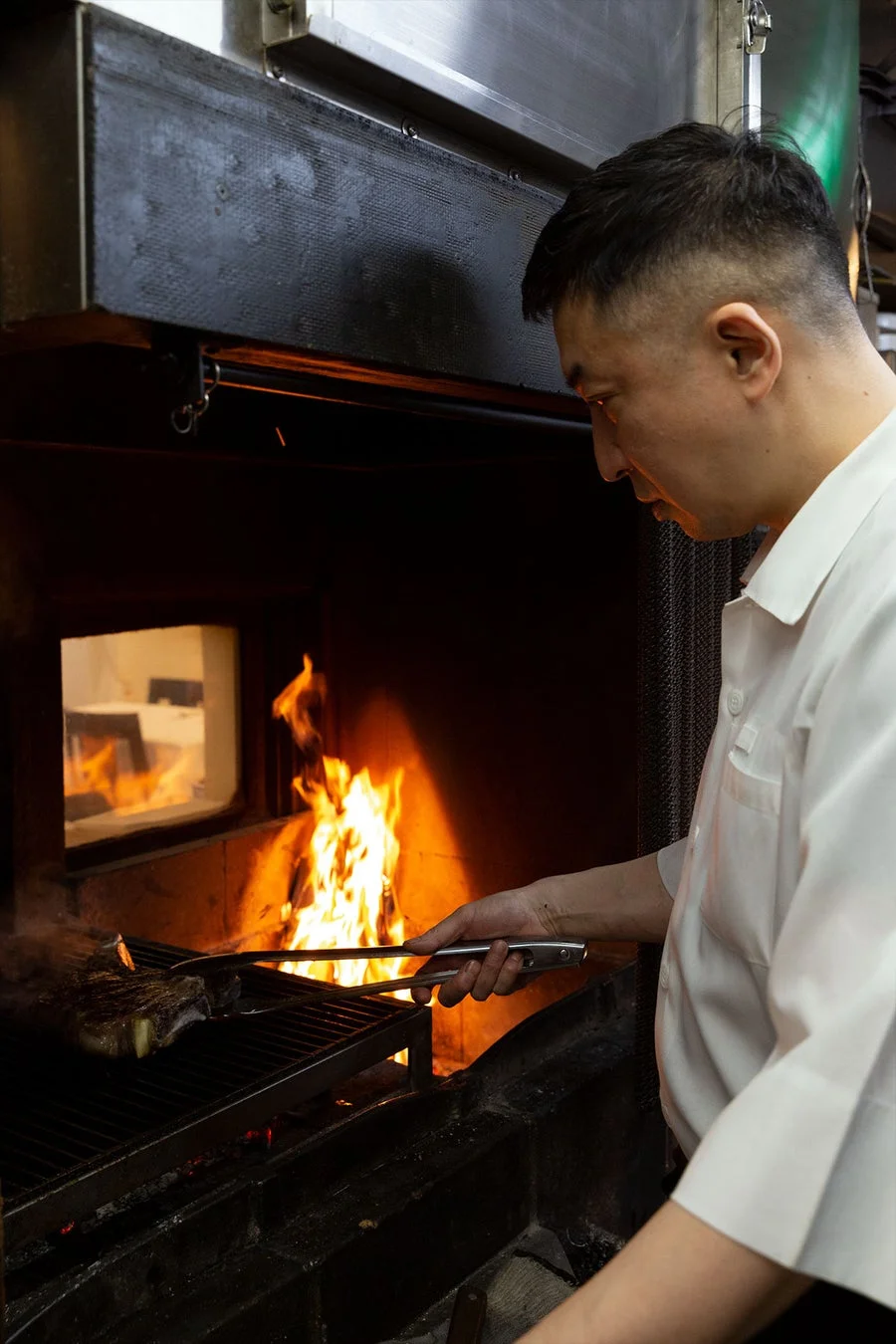 白いコックコートを着た料理人が、炎が燃え盛るグリルで肉を焼いています。トングを使い、丁寧に焼き加減を見ている様子が伺え、本格的な調理風景です。