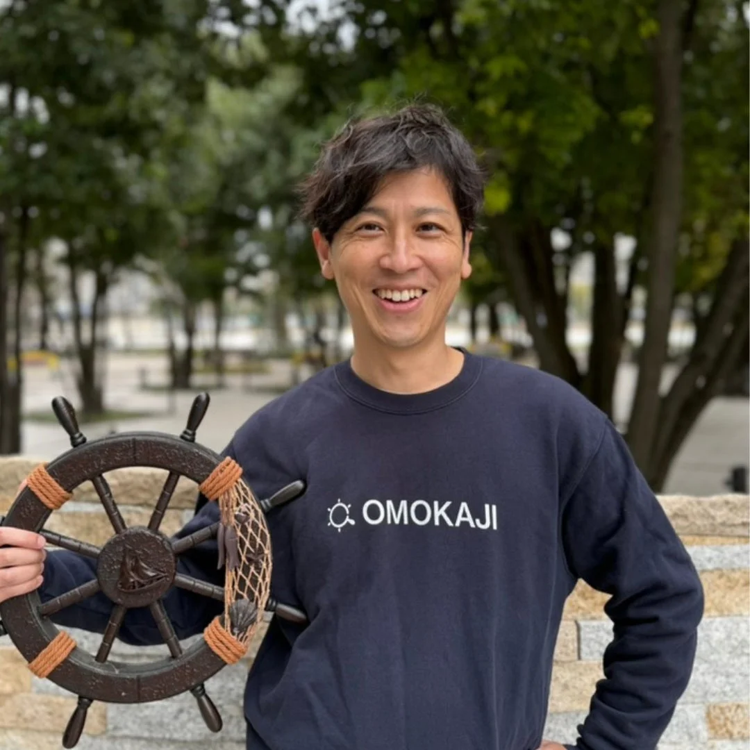 紺色のスウェットシャツを着た男性が、笑顔で船の舵の飾りを手に持っています。背景は木々が見える屋外の公園のような場所です。