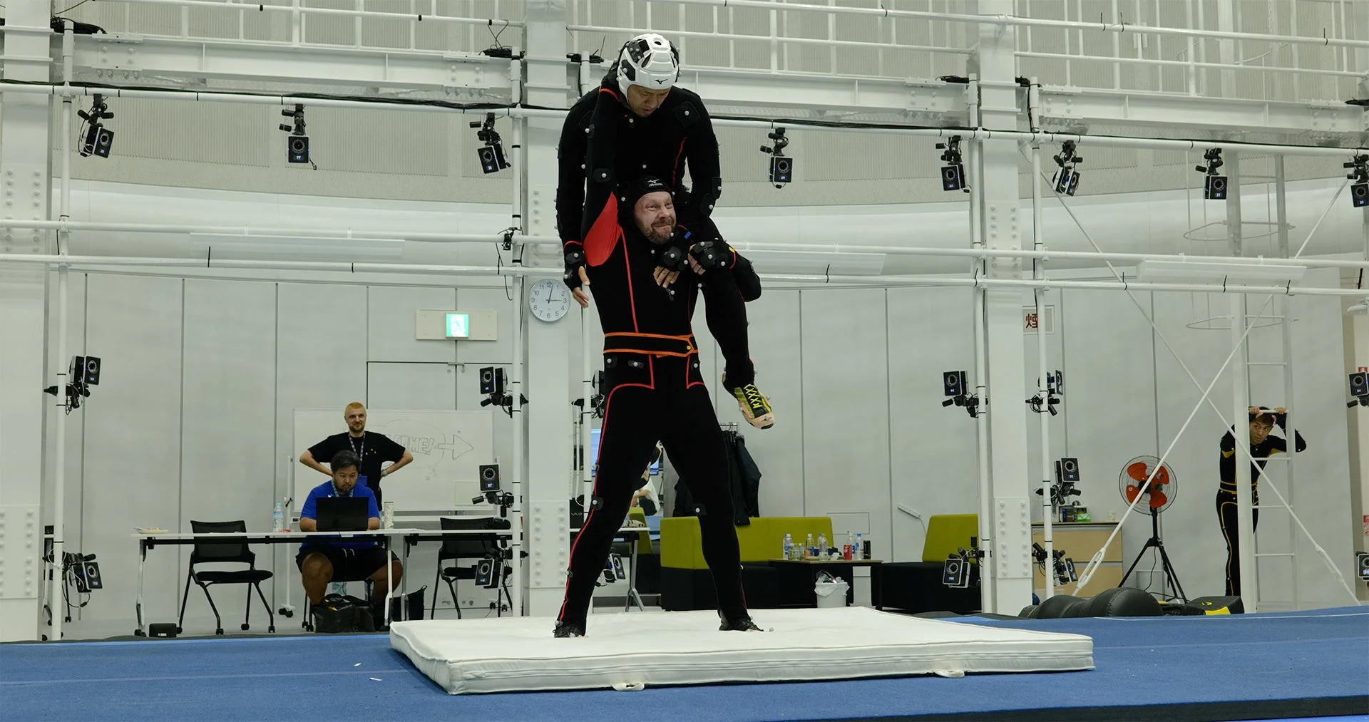 モーションキャプチャースタジオで、特殊なスーツを着た2人の男性がアクロバティックなポーズを取っている。一人がもう一人の肩に乗っており、周囲には多数のモーションキャプチャーカメラが設置されている。