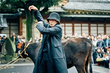 黒いコートと帽子を身につけた男性が牛の横でポーズをとっているパフォーマンスの様子