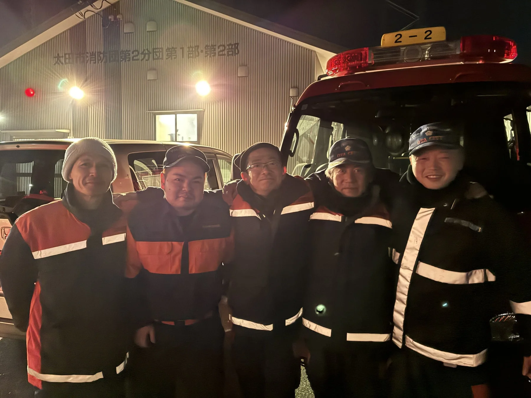 夜間、太田市消防団の建物と消防車の前で、5人の男性消防団員が制服姿で肩を組み、笑顔で記念撮影をしている様子です。