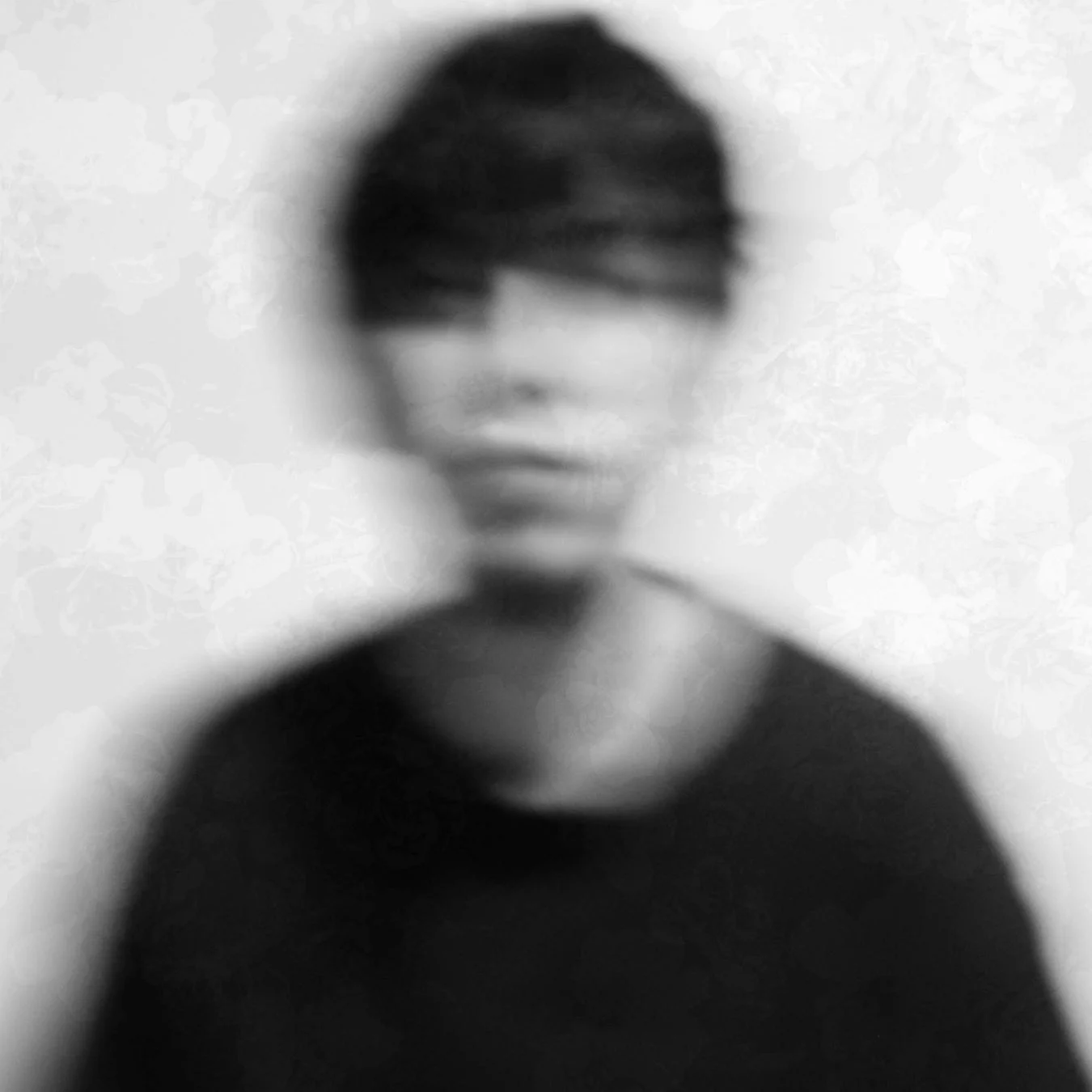 人物の顔がぼかされ、抽象的な表現がされた白黒のポートレートです。背景にはかすかな模様が見え、全体的に不明瞭で神秘的な雰囲気を醸し出しています。