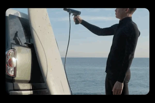 ウェットスーツを着た男性が、海辺で車に立てかけたサーフボードを携帯式の洗浄機で洗っている様子