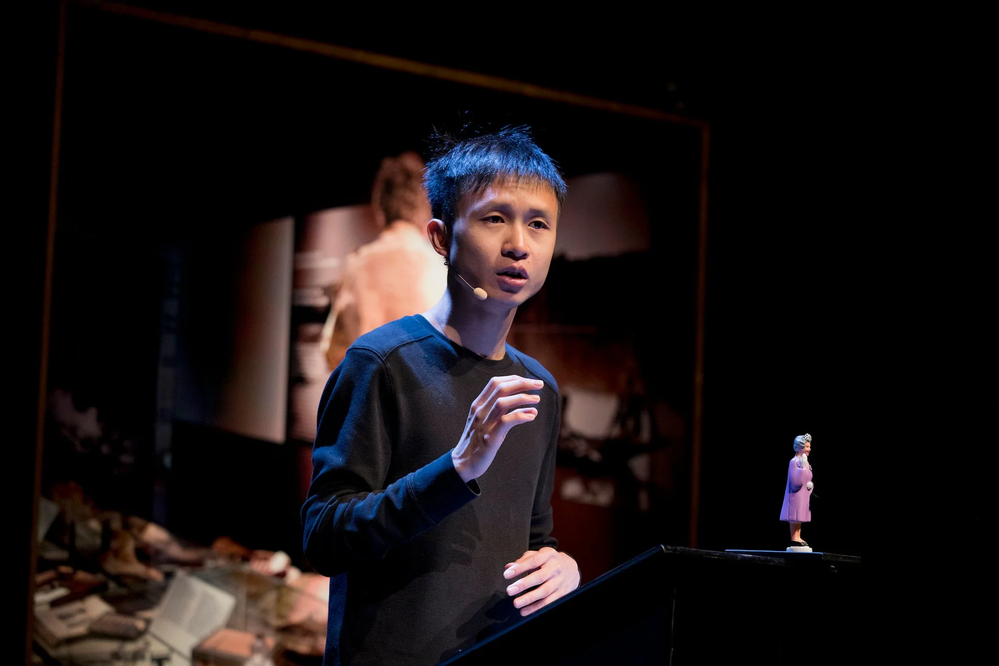 暗いステージ上で、ラベリアマイクを装着したアジア系の男性が熱心に話しています。