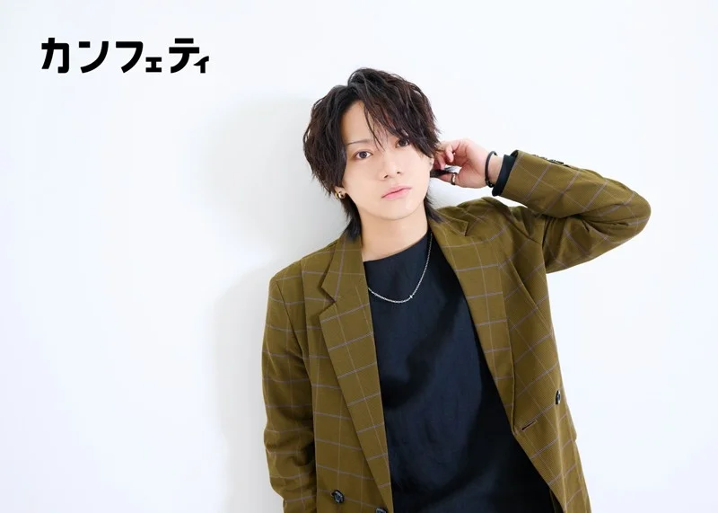 白い背景の前で、カーキ色のチェック柄ジャケットと黒いTシャツを着用した若々しい男性が、髪に手を当ててポーズをとっています。