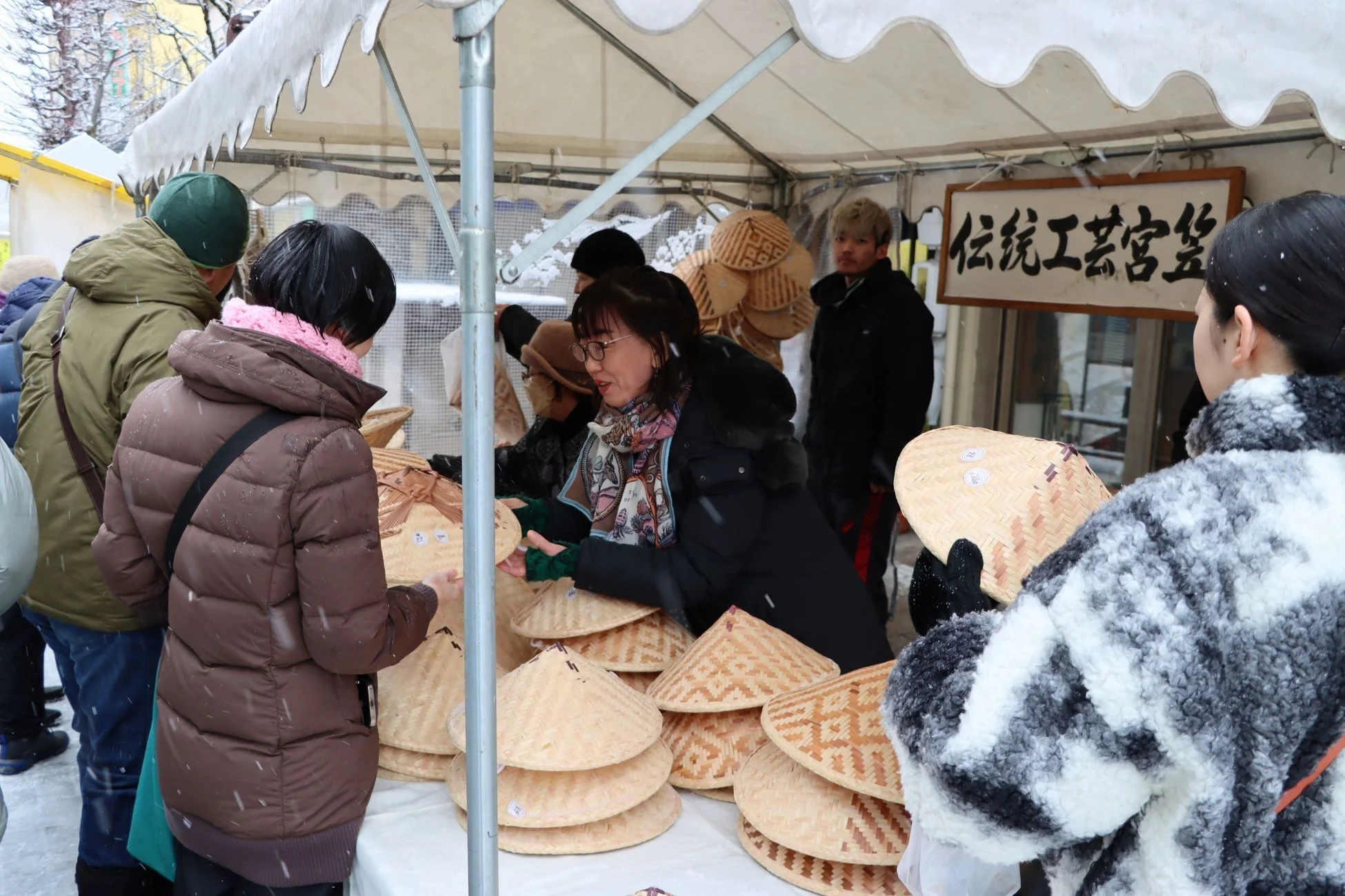 雪が降る冬の屋外マーケットで、人々が伝統工芸品である菅笠（すげがさ）を品定めしている様子。店には「伝統工芸菅笠」の看板が見える。