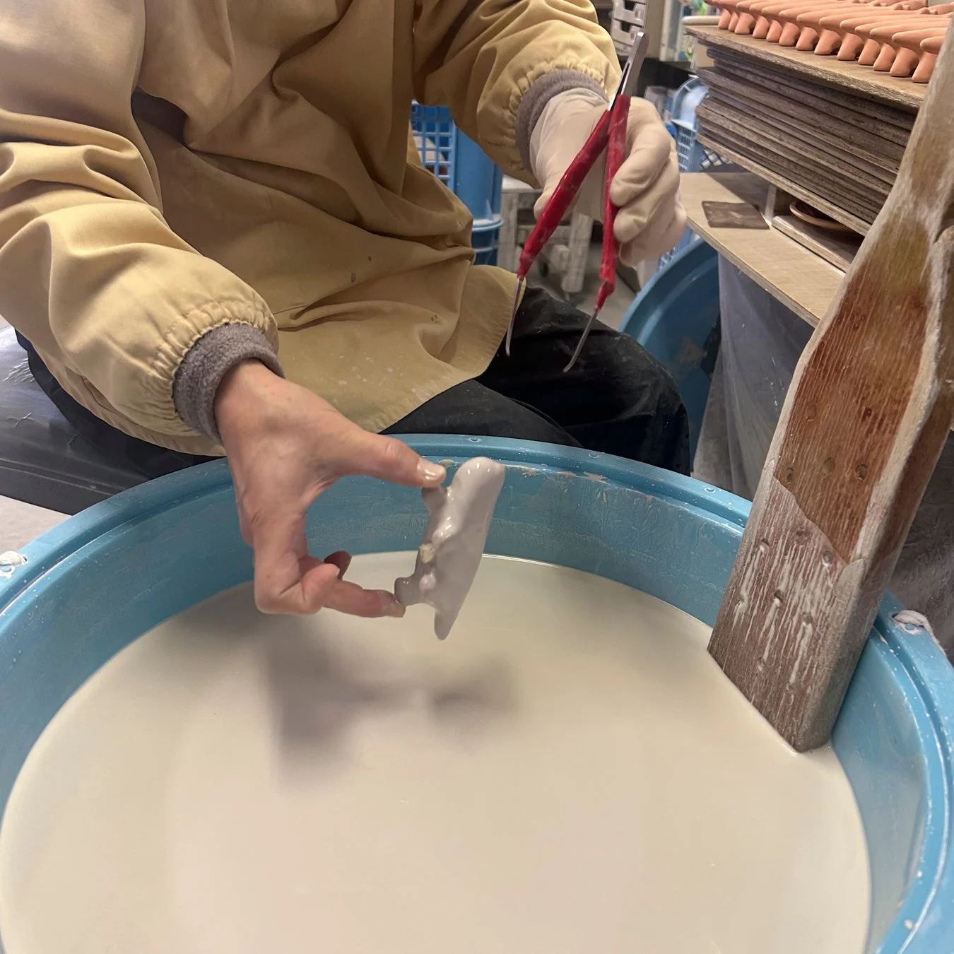 作業着を着た人が、青い桶に入った白い液体に素焼きの陶器を浸している様子です。陶器の製造工程で釉薬をかける作業をしていると思われ、手作業による工芸品の製作風景が伺えます。