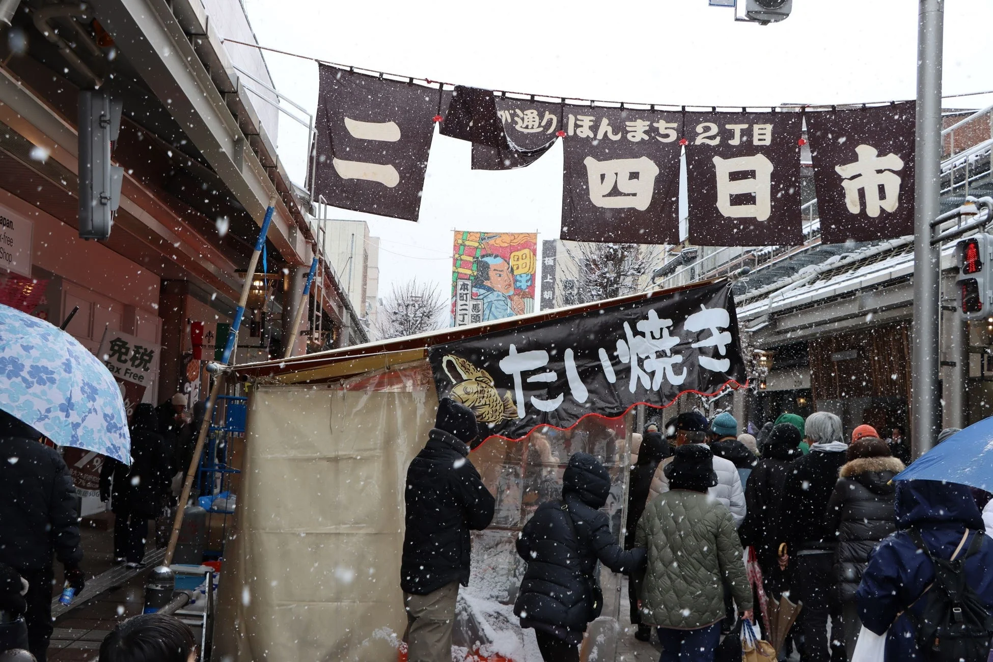雪が降る中、日本の商店街で多くの人々が歩いている様子。たい焼きの屋台や「四日市」と書かれた旗が見られ、賑やかな冬の市場の雰囲気が伝わってくる。