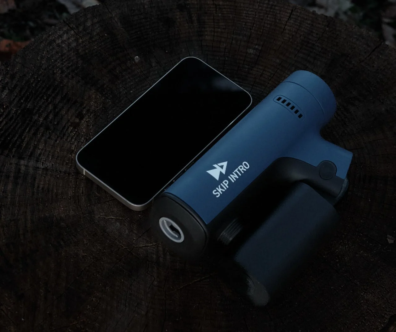 暗い木製の切り株の上に、画面が黒いスマートフォンと「SKIP INTRO」のロゴが入った青いガジェットが置かれています