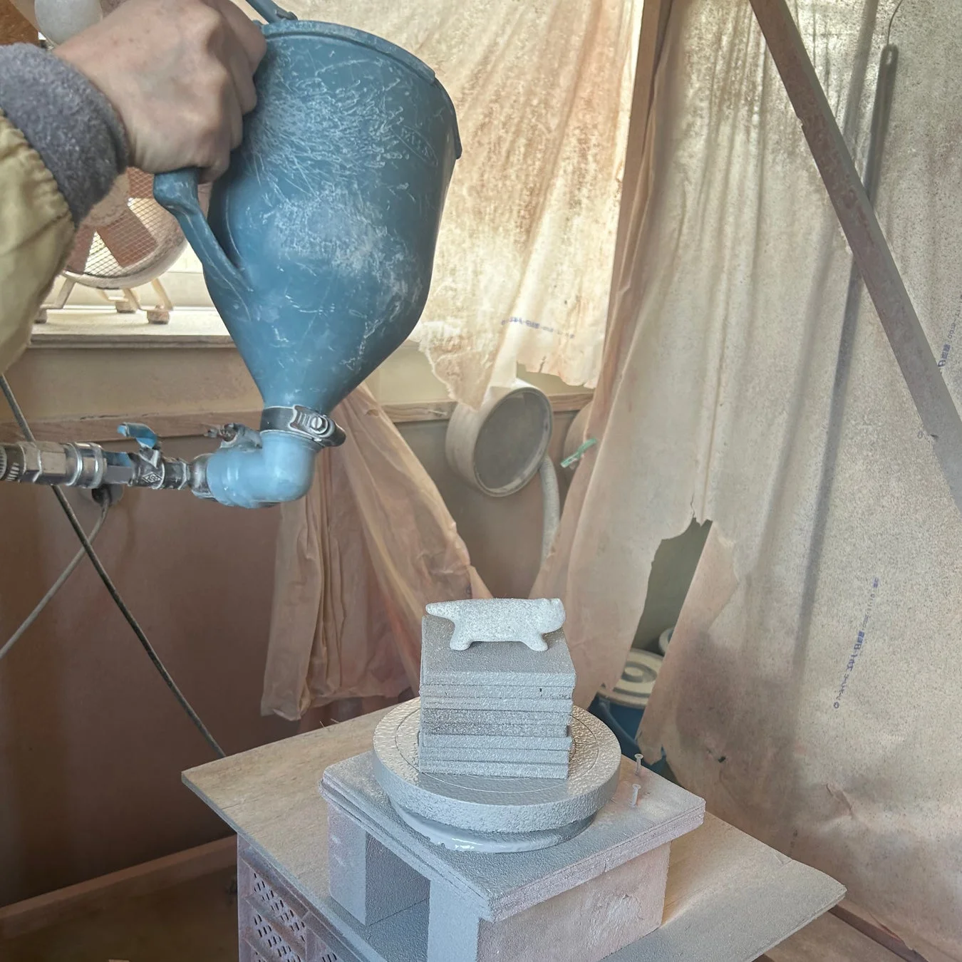 人物がスプレーガンを使い、積み重ねられた台座の上の猫の置物に粉体塗装のようなコーティングを施している作業風景です。工芸品や陶器の製造工程の一部と見られ、細やかな作業が進行中です。