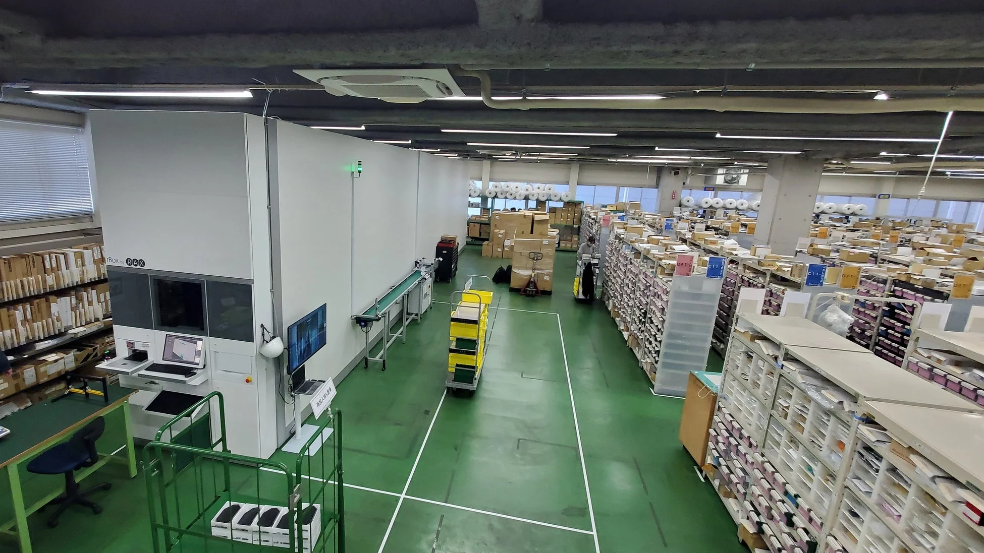 広々とした倉庫の内部で、自動倉庫システム「Box by RAX」やコンベアが稼働している様子です。多数の棚には商品が整然と並べられ、作業員がカートを使ってピッキング作業を行っており、効率的な物流プロセスがうかがえます。