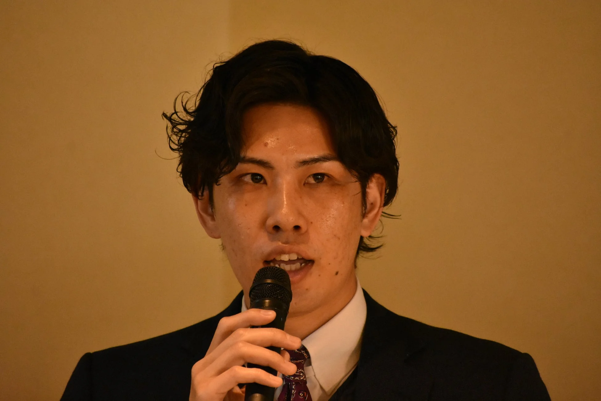 スーツを着た若い男性がマイクを持ち、熱心に話している様子を捉えたクローズアップ画像です。フォーマルなイベントやプレゼンテーションの場面を思わせます。