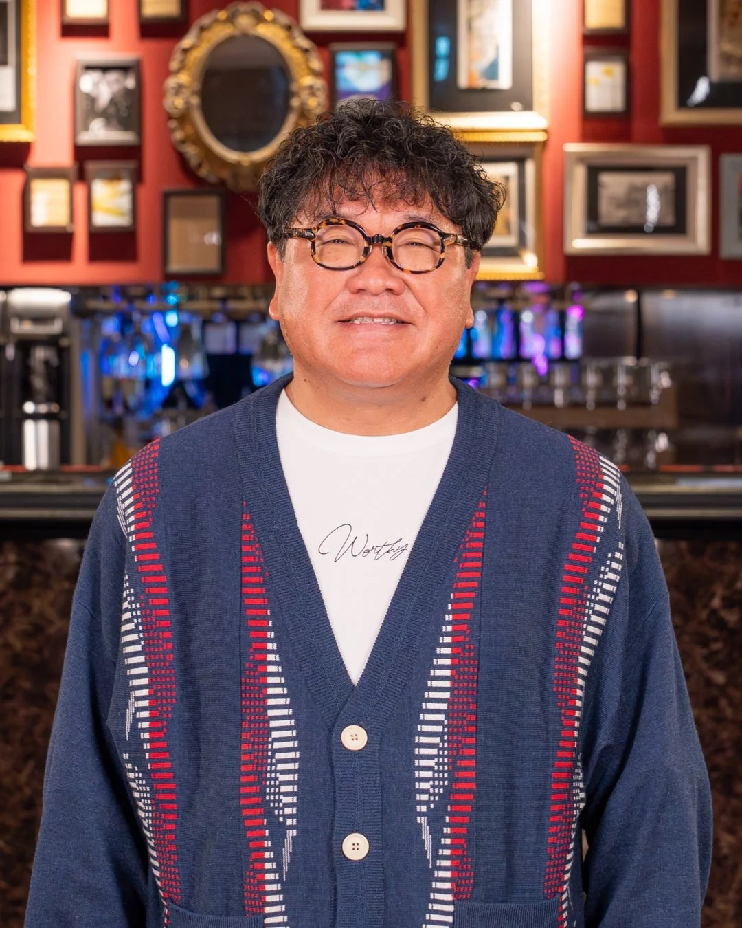 笑顔でカメラを見つめる中年の日本人男性のポートレート。メガネをかけ、紺色の柄入りカーディガンを着用しています。背景には赤い壁に額縁や鏡が飾られ、バーのような空間が見えます。