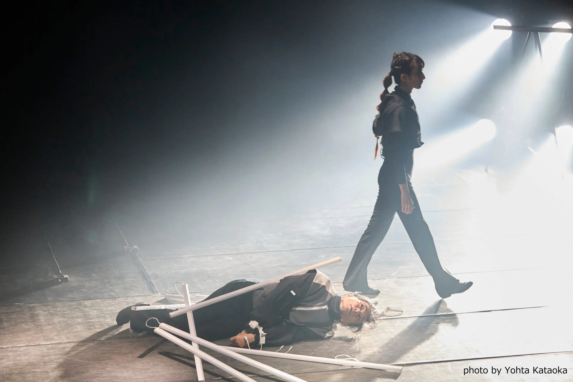 明るいスポットライトと霧が立ち込める舞台で、一人の人物が歩き、もう一人の人物が白い棒状のプロップに囲まれて横たわっています。動きと静止の対比が印象的な、演劇的またはパフォーマンスアートの一場面です。