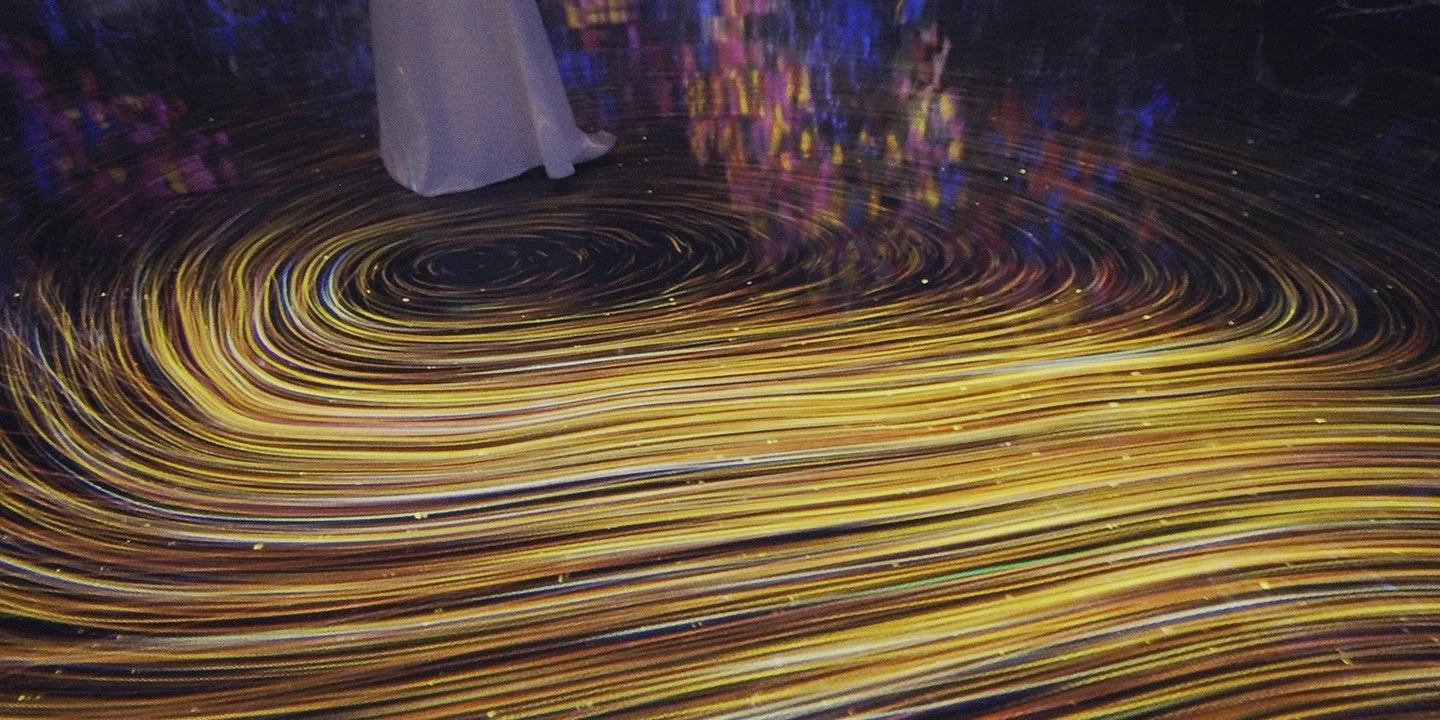 暗い空間の床に、渦巻くような金色の光のパターンが投影されており、その上に白いドレスをまとった人物の足元が立っています。