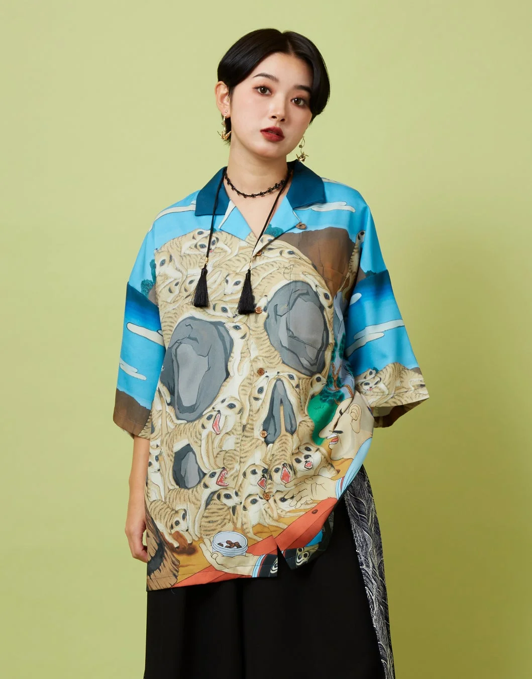 明るい緑の背景の前に立つ、ショートヘアの女性。ミーアキャットが群れるユニークな柄のカラフルなシャツを着用しており、全体的に個性的でアート性の高いファッションが特徴です。