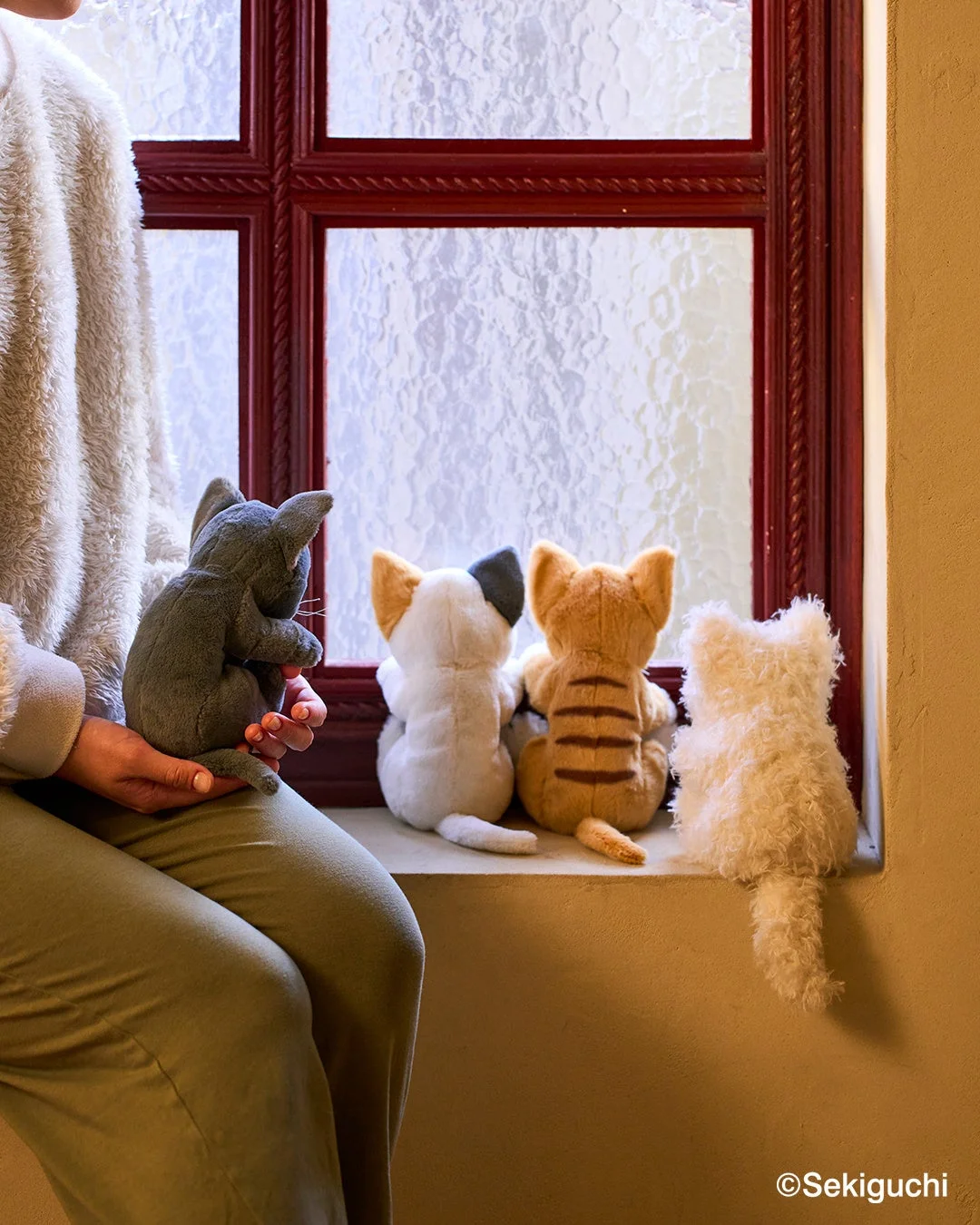 窓辺で人がグレーの猫のぬいぐるみを抱き、他の猫のぬいぐるみも眺めている様子