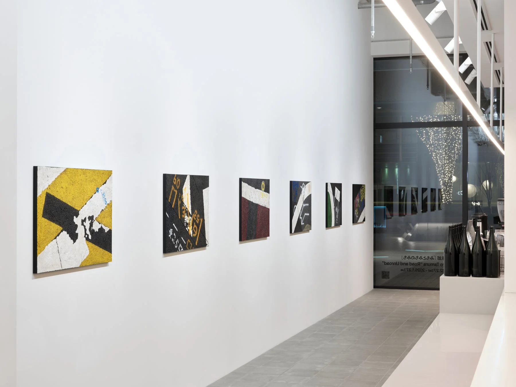 白い壁に抽象的な現代アート作品が展示されたギャラリーの風景。道路標識のような幾何学模様が特徴で、夜景の窓外にはイルミネーションが見える。右側にはボトルが並べられている。