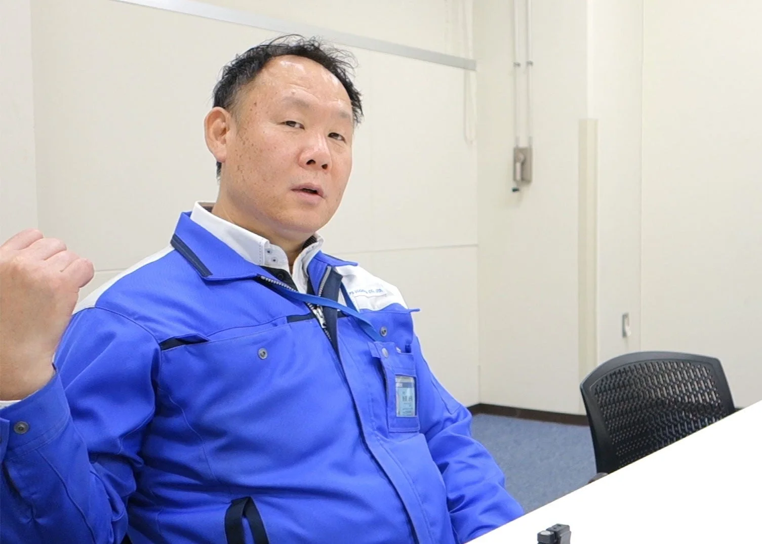 青と白の作業服を着た男性が、室内でテーブルに座っています。真剣な表情で、会議や打ち合わせ中の様子をうかがわせます。