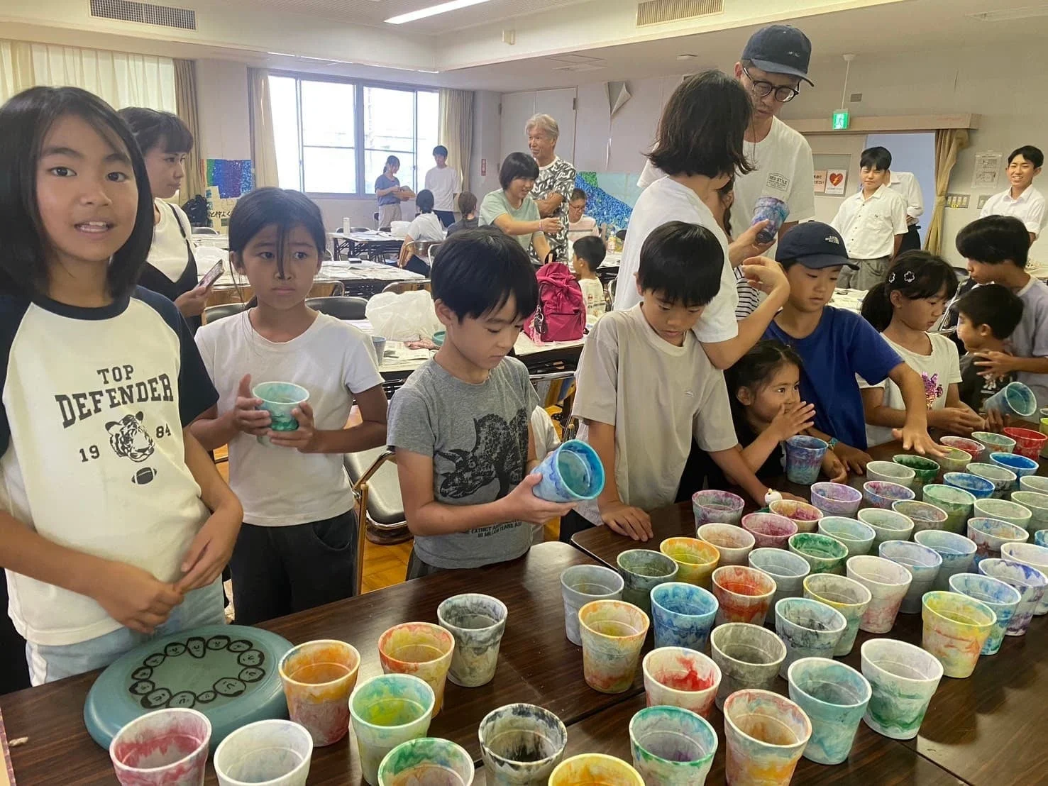 教室のような場所で、子供たちがカラフルなコップを並べ、絵付けなどのアート活動を楽しんでいる様子