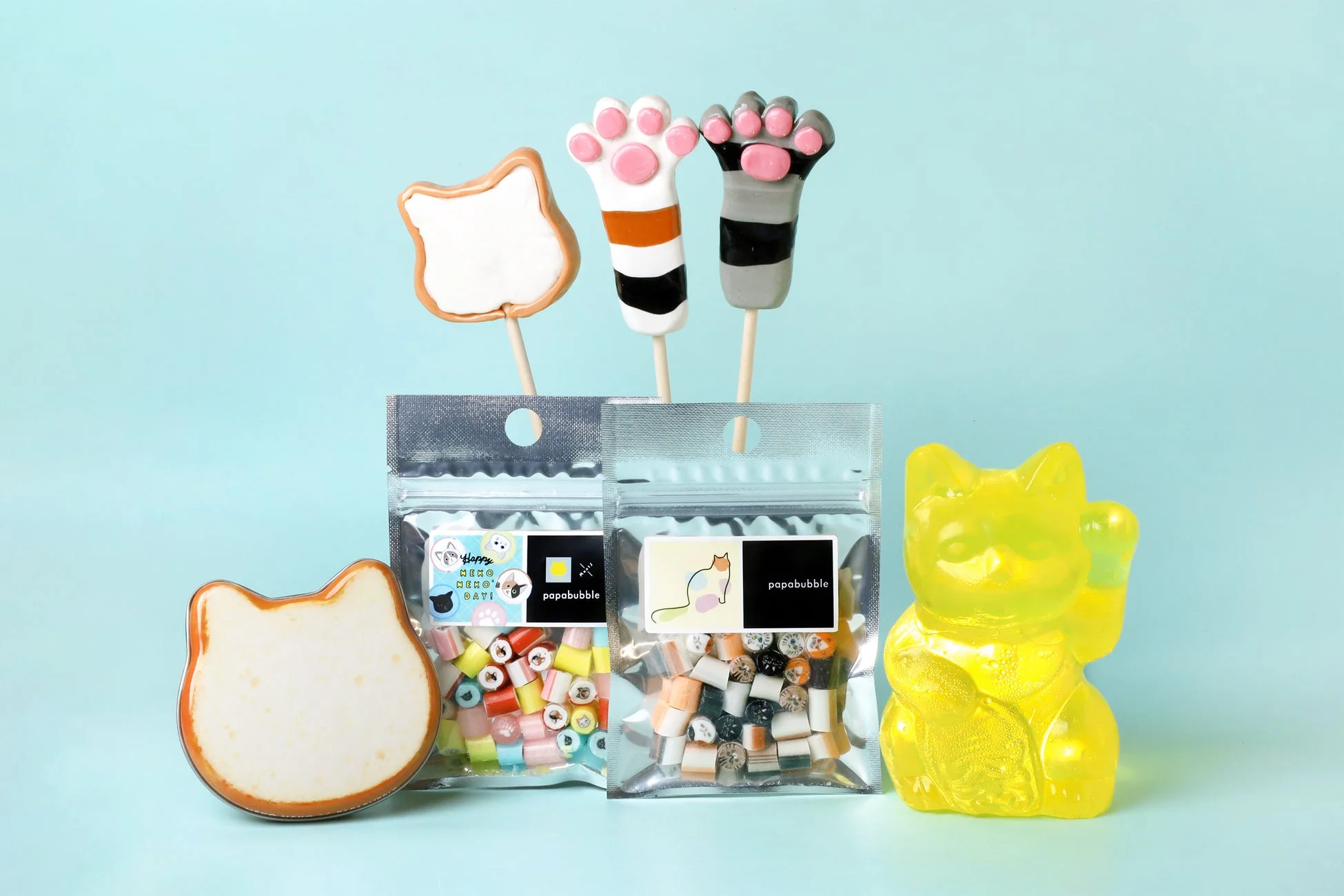 水色の背景に、猫の肉球ロリポップ、猫型クッキー、猫デザインのハードキャンディの袋、黄色の招き猫が並ぶ、可愛らしい猫テーマのスイーツの写真です。