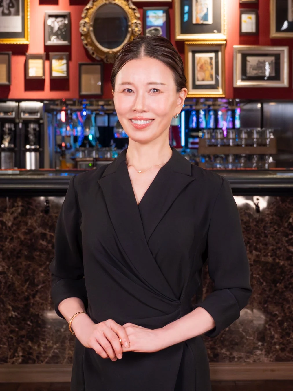 黒いドレスを着たアジア人女性が笑顔でカメラを見つめています。背景には額縁に入った絵画や鏡が飾られた、高級感のあるバーまたはレストランのような空間が広がっています。