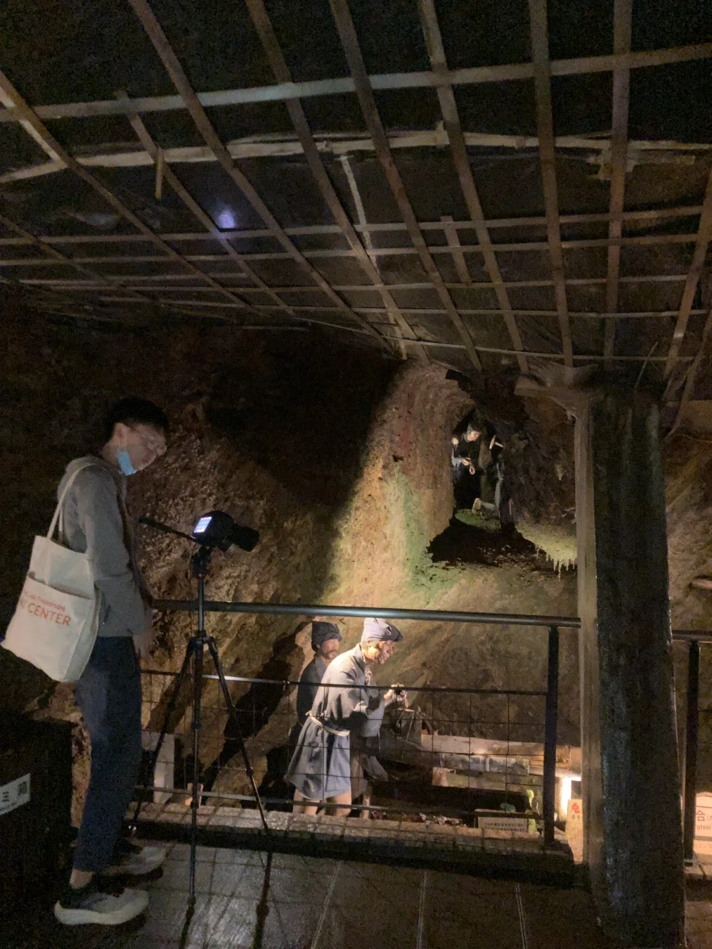 地下の坑道内で、マスクを着用した観光客が三脚を立てており、奥には採掘作業を行う様子の人形が展示されています。