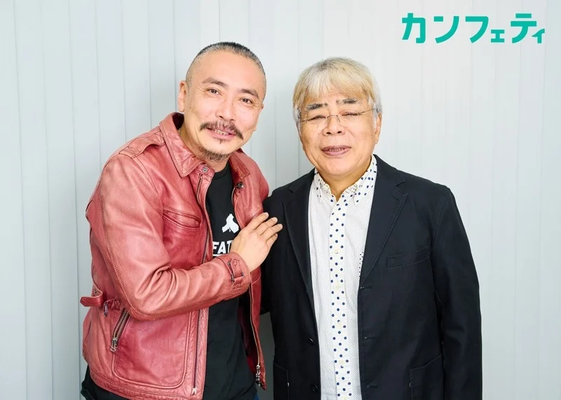 赤いレザージャケットを着た男性と、黒いジャケットに水玉シャツを着た男性が肩を組み、笑顔で並んでいます。