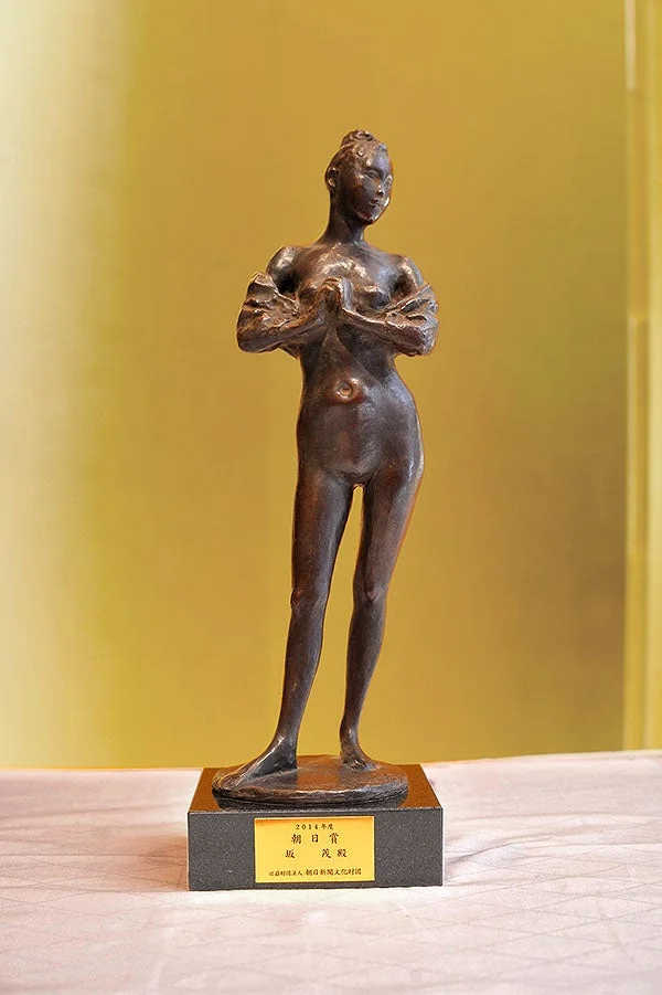 黄金色の背景を前に、台座に立つ女性のブロンズ像が写されています。台座には「2014年度 朝日賞 坂茂 殿」と記された金色のプレートがあり、朝日賞のトロフィーであることがわかります。
