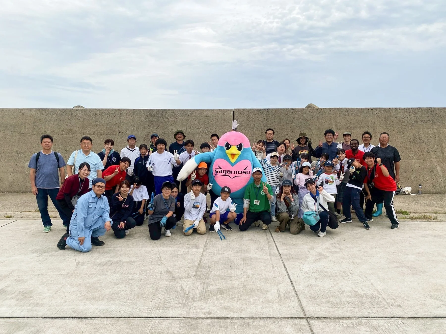 青とピンクの鳥の大きなマスコットを中心に、子供から大人まで多くの人々が集合写真を撮っている様子