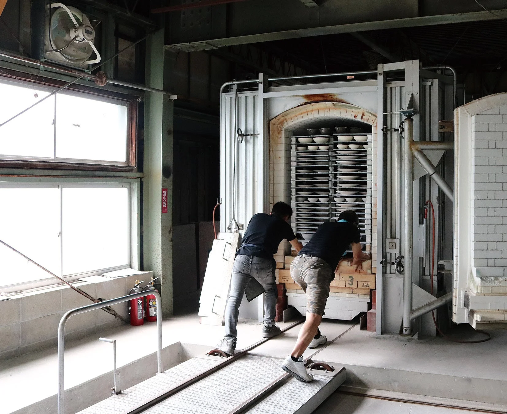 二人の男性が大量の陶器を積んだ台車を大きな窯の中に押し入れている様子。工場のような場所で、陶芸品の焼成準備をしている場面です。